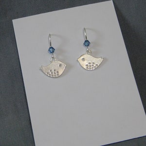May include: A pair of silver bird earrings, each with a small blue crystal bead. The bird-shaped charms feature a dotted wing design and a small circle for the eye. The earrings are presented on a white card.