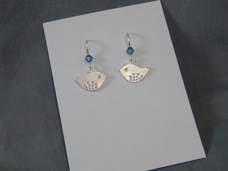 May include: A pair of silver bird earrings, each with a small blue crystal bead. The bird-shaped charms feature a dotted wing design and a small circle for the eye. The earrings are presented on a white card.