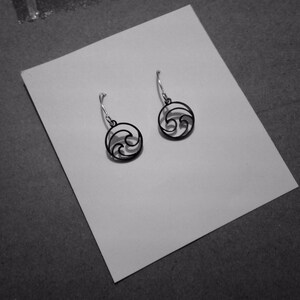 May include: A pair of black wave-shaped earrings with silver-colored hooks. The earrings are displayed on a white card against a dark gray background. The earrings have a circular design with a wave pattern.