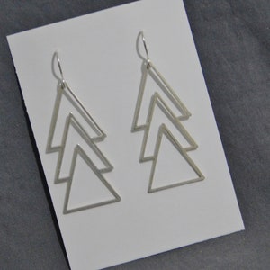 May include: Silver-toned geometric earrings featuring a layered triangle design. Each earring has three descending triangles, creating a modern, minimalist aesthetic. The earrings are displayed on a white card against a gray background.