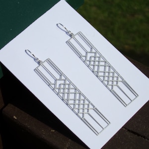 May include: A pair of silver geometric drop earrings. Each earring has a long rectangular shape with an open, lattice design. The earrings are presented on a white surface, with a green box and a brown surface in the background.