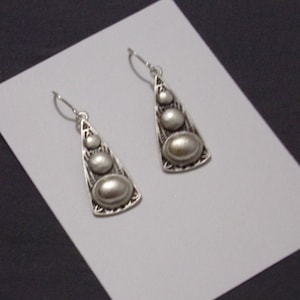 May include: A pair of silver-coloured earrings. Each earring features a triangular design with three graduated, oval-shaped embellishments. The earrings have a decorative filigree pattern around the edges and a hook closure.