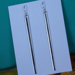 May include: A pair of silver-colored, linear dangle earrings. Each earring features a long, rectangular bar suspended from a curved ear wire. The earrings are displayed on a white card against a teal background, showcasing a minimalist jewelry design.