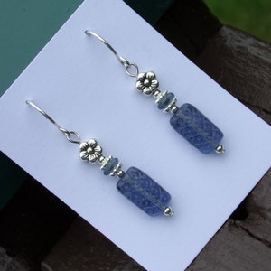 May include: A pair of dangle earrings with a floral design. The earrings feature blue rectangular beads with a carved pattern, silver-colored accents, and a flower-shaped top. The earrings are displayed on a white card.