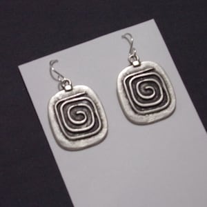 May include: A pair of silver-toned earrings. Each earring features a rectangular pendant with a raised spiral design. The earrings have a simple hook closure. The background is a light gray card against a dark gray surface.
