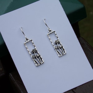 May include: Silver-toned mushroom earrings. Each earring features a rectangular frame with a textured border, and three mushroom figures. The earrings have a hook closure. The earrings are displayed on a white surface.