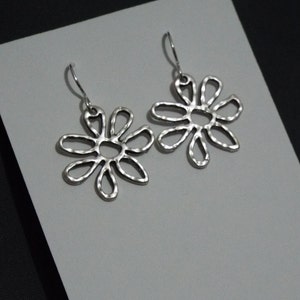 May include: A pair of silver-coloured flower-shaped earrings. Each earring has a daisy-like design with open petals and a hammered texture. The earrings hang from simple ear wires, set against a grey and black background.