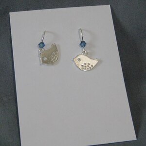 May include: A pair of silver bird earrings. Each earring features a bird-shaped charm with a small blue crystal accent. The birds have a textured design on their bodies and are suspended from silver hooks.
