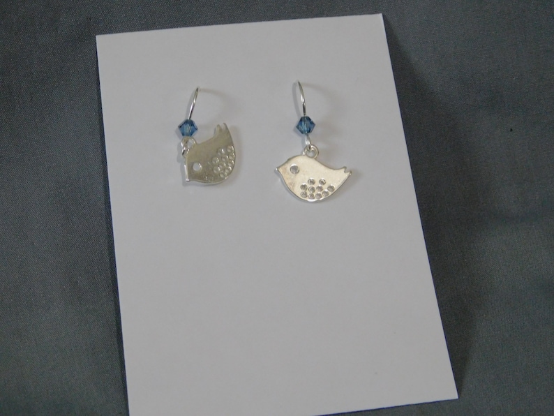 May include: A pair of silver bird earrings. Each earring features a bird-shaped charm with a small blue crystal accent. The birds have a textured design on their bodies and are suspended from silver hooks.