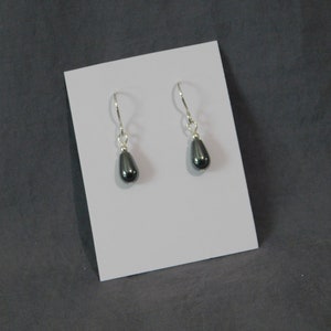 May include: A pair of drop earrings with dark gray, teardrop-shaped beads. The earrings hang from silver-colored, curved ear wires. The earrings are displayed on a white card against a gray background.