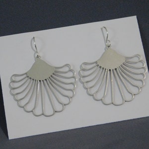 May include: A pair of silver fan-shaped earrings. Each earring has a solid, semi-circular top and a series of curved, radiating lines. The earrings are displayed on a white surface against a grey background.