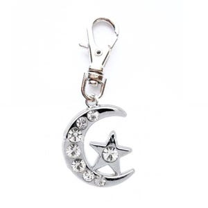 May include: Silver-colored keychain charm featuring a crescent moon and a star, both adorned with clear rhinestones. The charm is attached to a silver-tone metal clasp. The moon has seven rhinestones.
