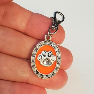 May include: Silver-colored pet tag with a paw print design. The tag has an orange background and is surrounded by a border of small, clear crystals. It includes a silver clasp for easy attachment to a collar or leash.