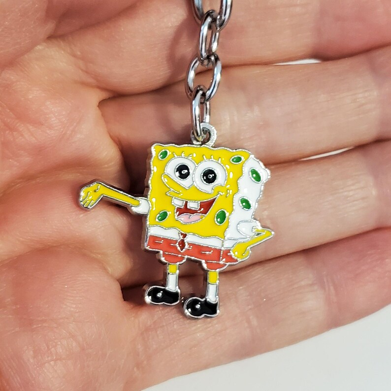May include: A yellow cartoon character keychain with a red and white outfit. The character has a big smile and is holding out its arm. The keychain is made of metal and has a silver chain.