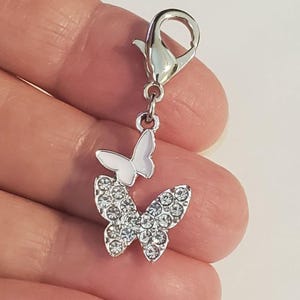 May include: A silver-toned charm with two butterfly pendants. The larger butterfly is encrusted with clear crystals, while the smaller one is white. The charm has a lobster clasp for easy attachment to a bracelet or necklace.