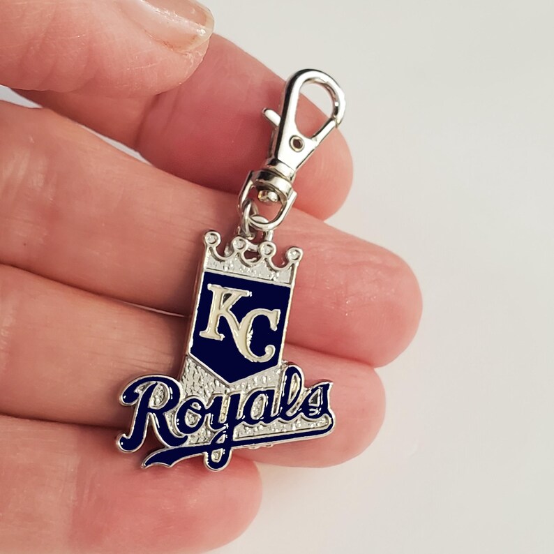 May include: A silver-toned keychain charm featuring the Kansas City Royals logo. The charm has a navy blue and white design with the letters "KC" inside a crown, and the word "Royals" in script. It includes a lobster clasp.