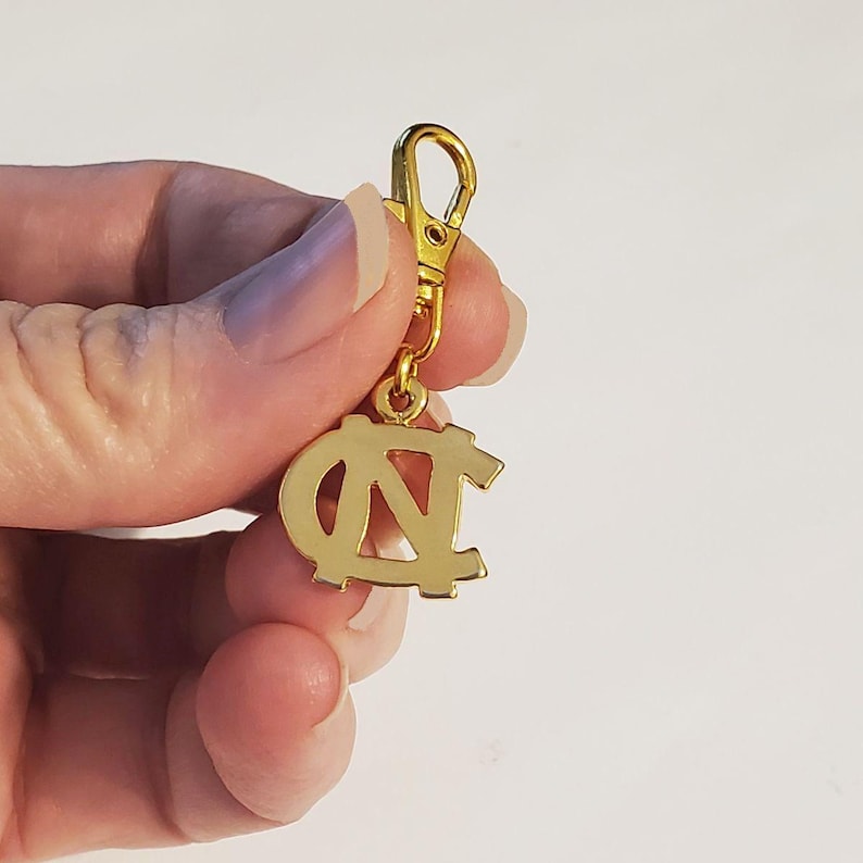 Op de afbeelding: Goudkleurige bedel met een sluiting, met het in elkaar grijpende "NC"-logo. De bedel is een helder, glanzend goud en lijkt van metaal te zijn. De sluiting is ook goudkleurig en is aan de bovenkant van de bedel bevestigd.