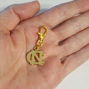 Op de afbeelding: Een goudkleurige bedel met een karabijnhaak. De bedel heeft de in elkaar grijpende letters "NC" in een gestileerd ontwerp. De bedel is waarschijnlijk gemaakt van metaal en is ontworpen om aan een sleutelhanger of ander accessoire te worden bevestigd.
