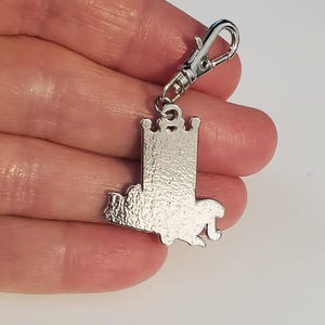 May include: A silver-colored charm with a castle design and a lobster clasp. The charm features a detailed castle silhouette with a crown on top. The clasp is attached to the top of the charm, allowing it to be easily attached to a keychain or bag.