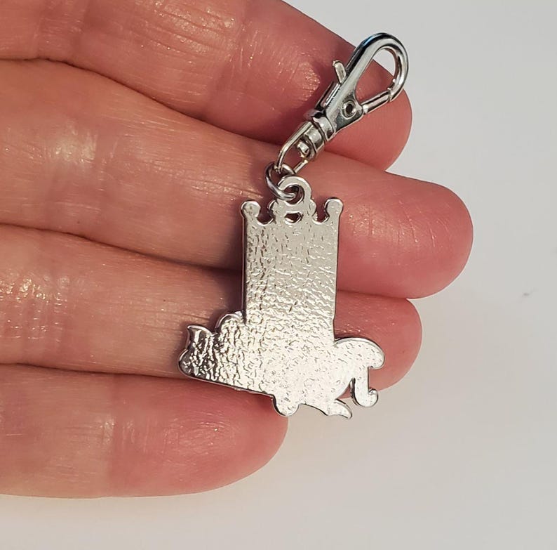 May include: A silver-colored charm with a castle design and a lobster clasp. The charm features a detailed castle silhouette with a crown on top. The clasp is attached to the top of the charm, allowing it to be easily attached to a keychain or bag.