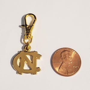 Op de afbeelding: Een goudkleurige bedel met een karabijnhaak, met de in elkaar grijpende letters "NC". Een munt ligt naast de bedel ter vergelijking van de grootte. De bedel is waarschijnlijk een sleutelhanger of accessoire.