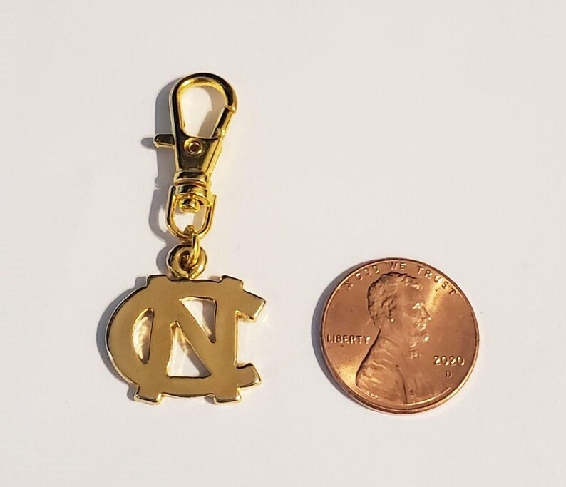 Op de afbeelding: Een goudkleurige bedel met een karabijnhaak, met de in elkaar grijpende letters "NC". Een munt ligt naast de bedel ter vergelijking van de grootte. De bedel is waarschijnlijk een sleutelhanger of accessoire.