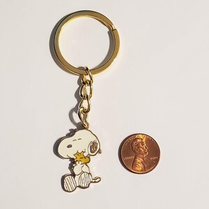 May include: A gold-colored keychain featuring a Snoopy character charm. The charm is white, yellow, and black, with a gold-colored chain and keyring. A penny is next to the keychain for size comparison.