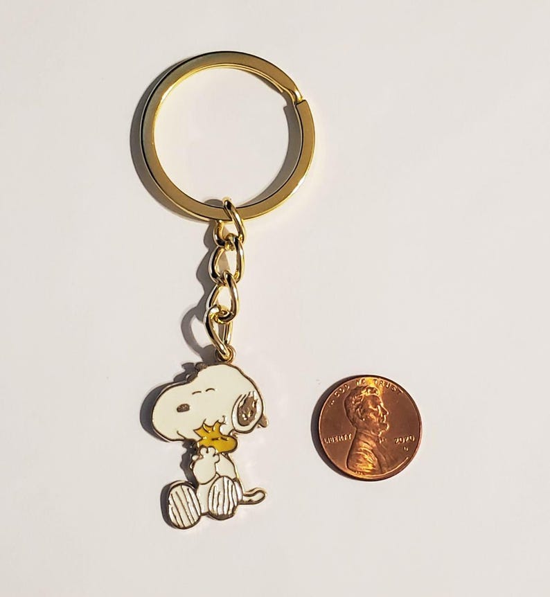 May include: A gold-colored keychain featuring a Snoopy character charm. The charm is white, yellow, and black, with a gold-colored chain and keyring. A penny is next to the keychain for size comparison.