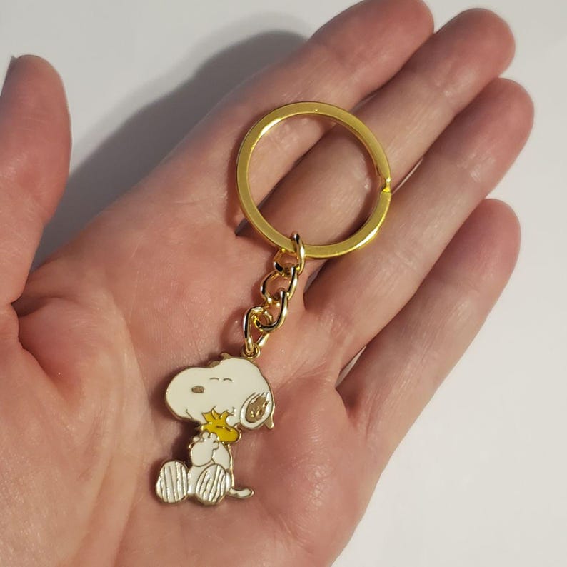 May include: A gold-colored keychain featuring a Snoopy charm. The charm depicts Snoopy in a seated position, with a yellow bird. The keychain has a gold ring and chain.
