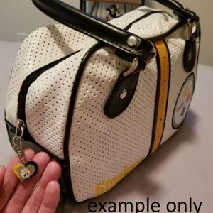 May include: White perforated handbag with black trim, handles, and zipper. The bag features yellow stripes and a team logo. A heart-shaped charm with team colors hangs from the zipper. The text "example only" is visible.