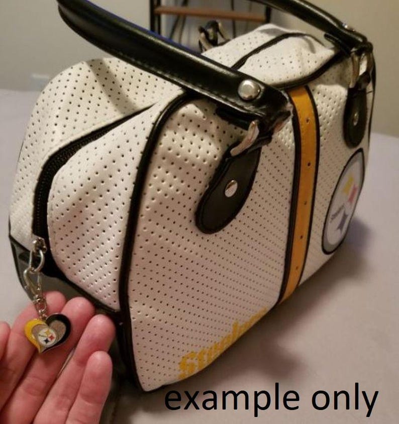 May include: White perforated handbag with black trim, handles, and zipper. The bag features yellow stripes and a team logo. A heart-shaped charm with team colors hangs from the zipper. The text "example only" is visible.