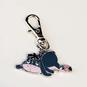 May include: A silver metal keychain featuring a cartoon character of Eeyore the donkey from Winnie the Pooh. Eeyore is lying down with his head resting on his front legs. He has a pink nose and a black and blue body.