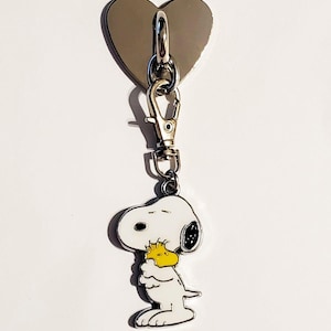 May include: A Snoopy keychain with a silver heart-shaped top and a clasp. The Snoopy charm is white with black accents and a yellow Woodstock. The keychain is made of metal and is attached to a silver clasp.