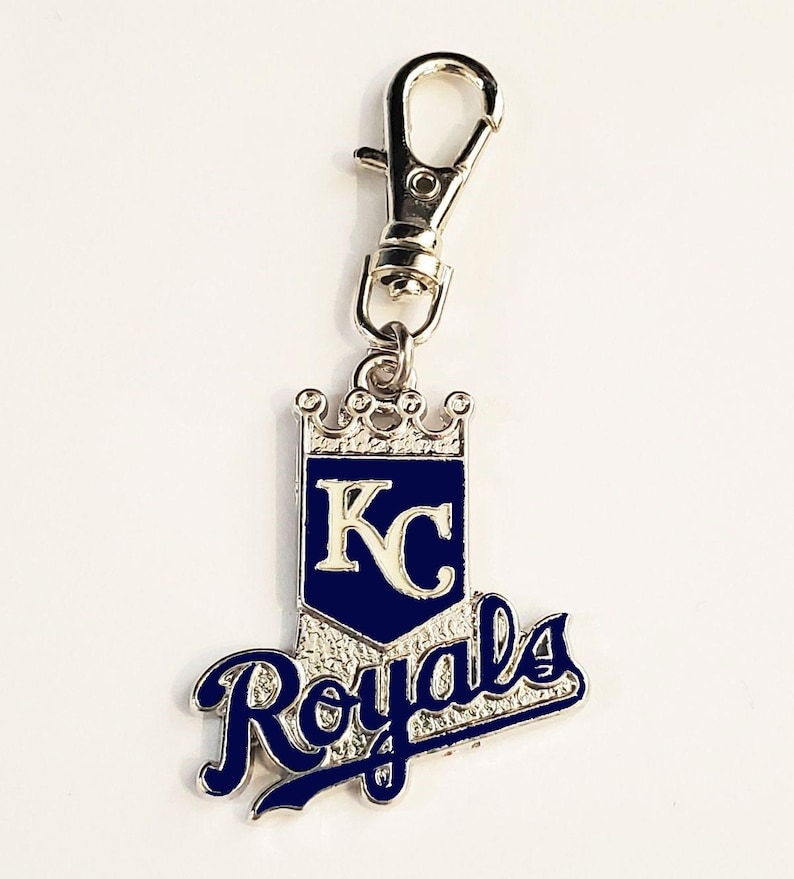 May include: A silver-toned keychain with a lobster clasp. The pendant features a blue and white design with the letters "KC" inside a shield, topped with a crown. The word "Royals" is written in cursive below.