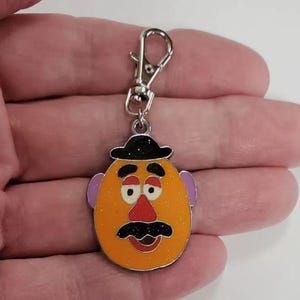 K&ouml;nnte beinhalten: Ein silberner Schl&uuml;sselanh&auml;nger mit einer Comicfigur mit braunem Hut, orangefarbenem Gesicht, lila Ohren, roter Nase und schwarzem Schnurrbart.