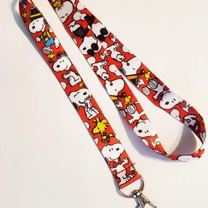 May include: A red lanyard featuring a repeating pattern of Snoopy and Woodstock characters in white, black, and yellow. The lanyard has a metal clasp at the end for attaching keys or an ID badge.