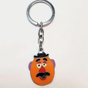 K&ouml;nnte beinhalten: Ein silberner Schl&uuml;sselanh&auml;nger mit einem Mr. Potato Head-Anh&auml;nger aus Zeichentrick. Der Anh&auml;nger ist orange mit einem schwarzen Hut, braunen Augenbrauen, lila Ohren, einer roten Nase und einem schwarzen Schnurrbart.