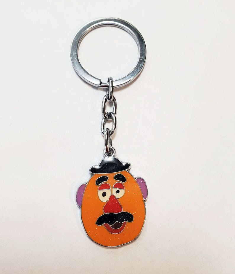 K&ouml;nnte beinhalten: Ein silberner Schl&uuml;sselanh&auml;nger mit einem Mr. Potato Head-Anh&auml;nger aus Zeichentrick. Der Anh&auml;nger ist orange mit einem schwarzen Hut, braunen Augenbrauen, lila Ohren, einer roten Nase und einem schwarzen Schnurrbart.