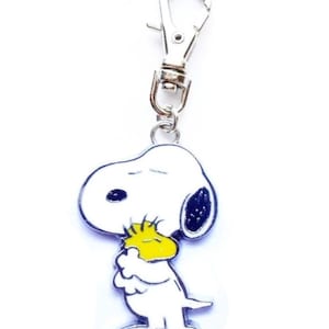 May include: A Snoopy keychain charm. The charm features a white Snoopy figure with black ears and a yellow bird on his chest. The charm is attached to a silver-colored metal clip.