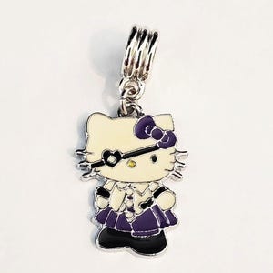 May include: A silver charm featuring a cartoon cat character with a cream-colored face, purple bow, and matching dress. The charm has a black heart-shaped eye mask and is suspended from a silver loop.