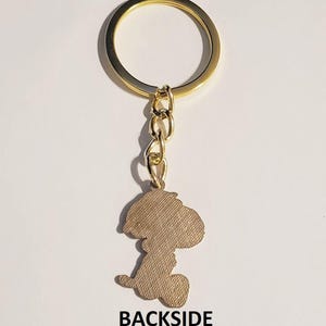May include: Gold-toned keychain featuring a Snoopy silhouette charm. The charm is a light brown color with a textured surface. The keychain includes a gold ring and chain, with the word "BACKSIDE" printed at the bottom.