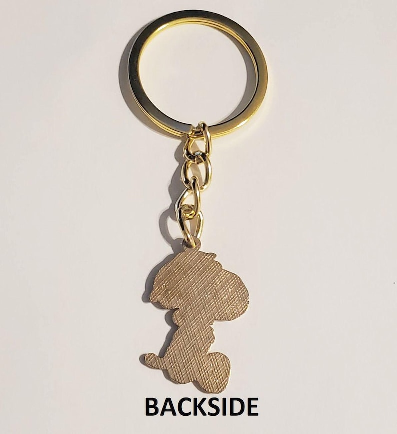 May include: Gold-toned keychain featuring a Snoopy silhouette charm. The charm is a light brown color with a textured surface. The keychain includes a gold ring and chain, with the word "BACKSIDE" printed at the bottom.