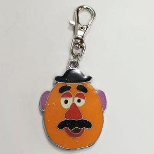 K&ouml;nnte beinhalten: Ein silberner Schl&uuml;sselanh&auml;nger mit einer Comicfigur mit schwarzem Hut, orangefarbenem Gesicht, schwarzem Schnurrbart und roter Nase. Die Augen der Figur sind geschlossen und der Schl&uuml;sselanh&auml;nger hat einen silbernen Clip.