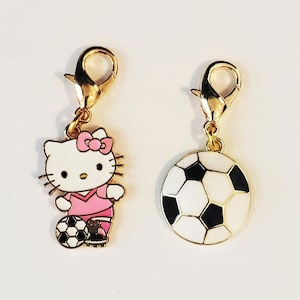 May include: Two gold-toned charm pendants. One features a cartoon cat in a pink soccer uniform with a soccer ball. The other is a soccer ball with black and white panels. Both charms have a lobster clasp.