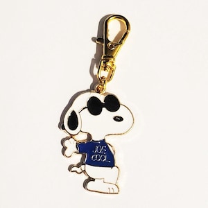 May include: A Snoopy keychain with a gold-colored clasp. The white Snoopy figure wears black sunglasses and a blue shirt with the text "JOE COOL". The keychain is a fun accessory.