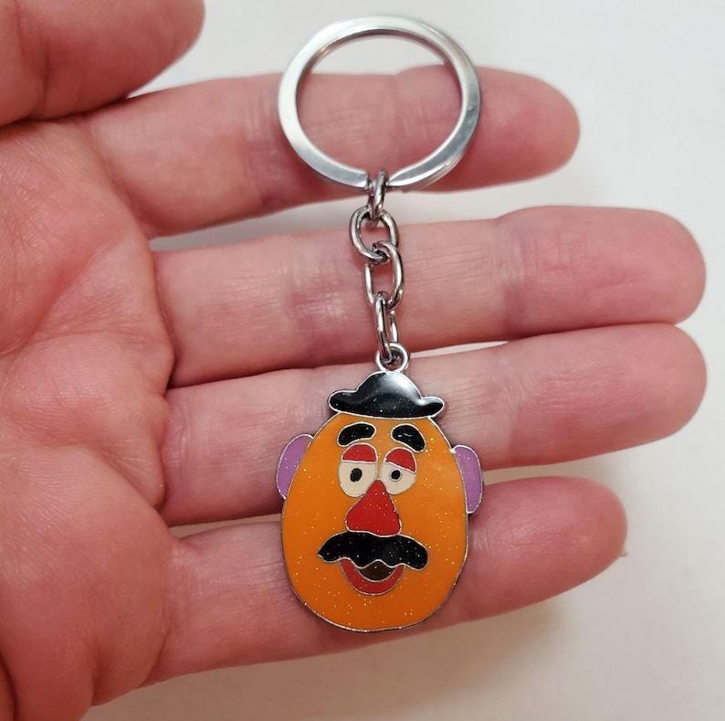K&ouml;nnte beinhalten: Ein silberner Schl&uuml;sselanh&auml;nger mit einem Mr. Potato Head-Anh&auml;nger. Der Anh&auml;nger ist orange mit einem schwarzen Hut, braunen Augenbrauen, einer roten Nase und einem schwarzen Schnurrbart.