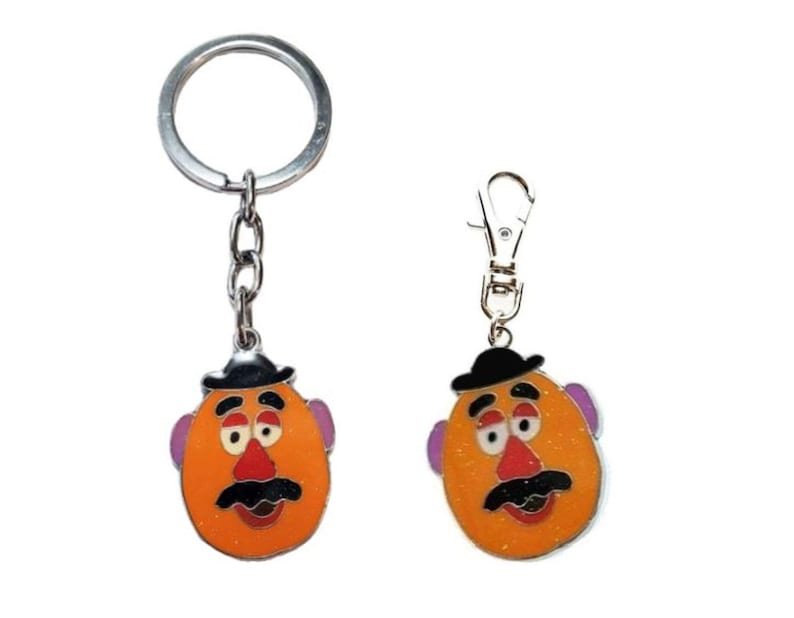 K&ouml;nnte beinhalten: Zwei Schl&uuml;sselanh&auml;nger aus Metall mit Mr. Potato Head. Der gr&ouml;&szlig;ere Schl&uuml;sselanh&auml;nger hat einen silbernen Ring und eine Kette. Der kleinere Schl&uuml;sselanh&auml;nger hat einen silbernen Karabinerhaken. Beide Schl&uuml;sselanh&auml;nger zeigen ein Cartoon-Bild von Mr. Potato Head mit einem schwarzen Hut, orangefarbener Haut, lila Ohren, einer roten Nase und einem schwarzen Schnurrbart.
