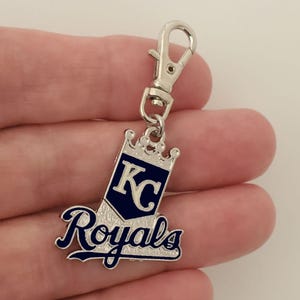 May include: A silver-colored keychain charm with a lobster clasp. The charm features the words "Royals" in a script font and the letters "KC" inside a navy blue pennant shape, topped with a crown.