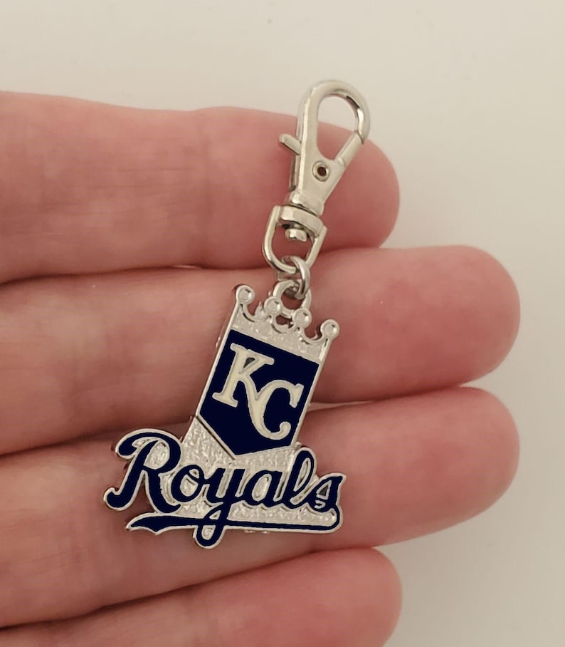 May include: A silver-colored keychain charm with a lobster clasp. The charm features the words "Royals" in a script font and the letters "KC" inside a navy blue pennant shape, topped with a crown.