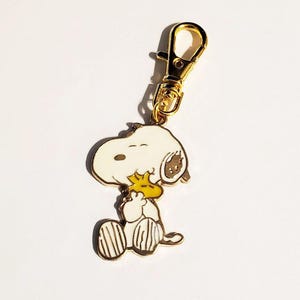 May include: A Snoopy keychain with a gold-colored clasp. The charm features a white Snoopy character with black and brown accents, holding a yellow bird. The keychain is made of metal and has a gold-colored finish.